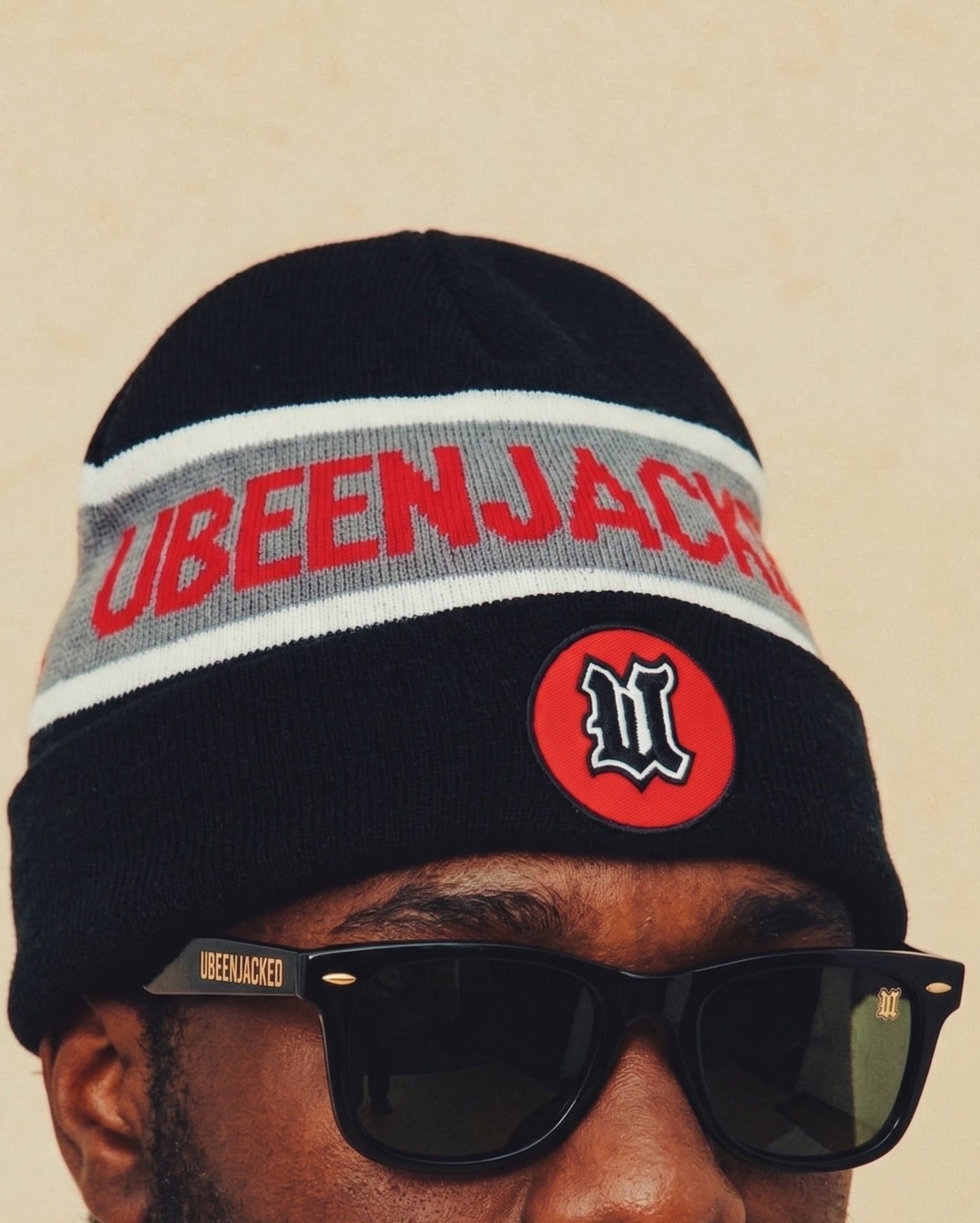 A close-up of a person wearing a black knit beanie with a gray stripe, red lettering that reads 'UBEENJACKER', and a red circular logo patch, along with black sunglasses against a beige background.