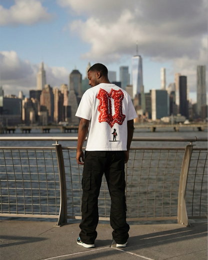 Person wearing a white t-shirt with red and black design, standing by a waterfront with a city skyline in the background.