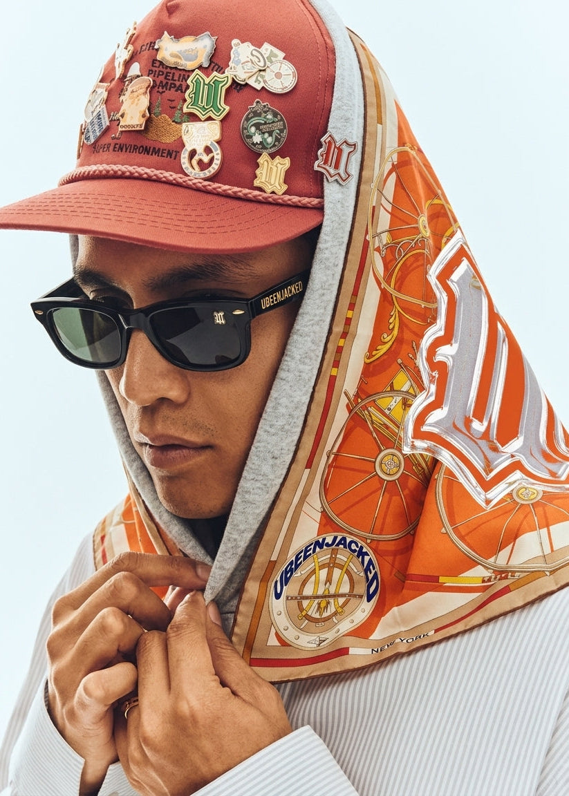 A close-up lifestyle shot of a person wearing a red baseball cap covered in pins, black sunglasses, and a gray hoodie with an orange and beige patterned scarf draped over their head and shoulders.