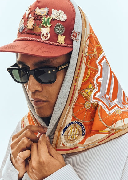 A close-up lifestyle shot of a person wearing a red baseball cap covered in pins, black sunglasses, and a gray hoodie with an orange and beige patterned scarf draped over their head and shoulders.